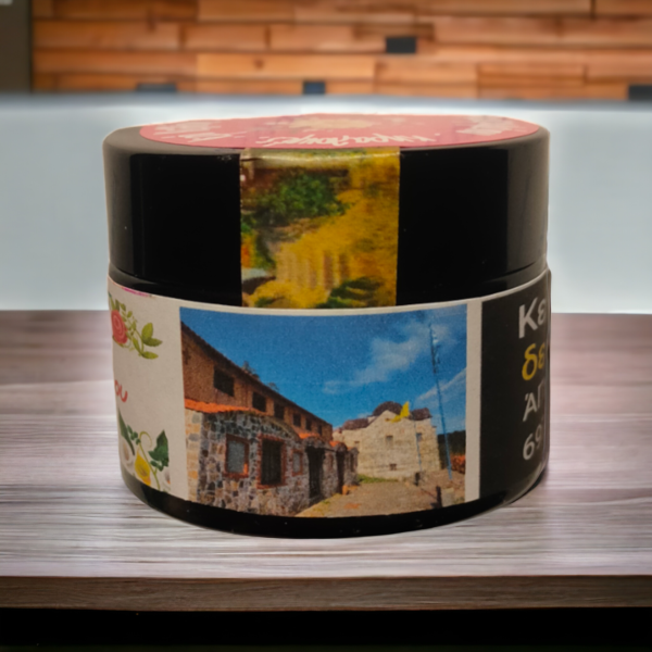 A small black container with a label featuring images of a picturesque stone building under a bright blue sky and a scenic garden. The label also includes text in Greek. The container is placed on a wooden surface with a blurred background. Ένα μικρό μαύρο δοχείο με ετικέτα που απεικονίζει εικόνες από ένα γραφικό πέτρινο κτίριο κάτω από έναν φωτεινό γαλάζιο ουρανό και έναν γραφικό κήπο. Η ετικέτα περιλαμβάνει επίσης κείμενο στα Ελληνικά. Το δοχείο είναι τοποθετημένο σε ξύλινη επιφάνεια με θολό φόντο.